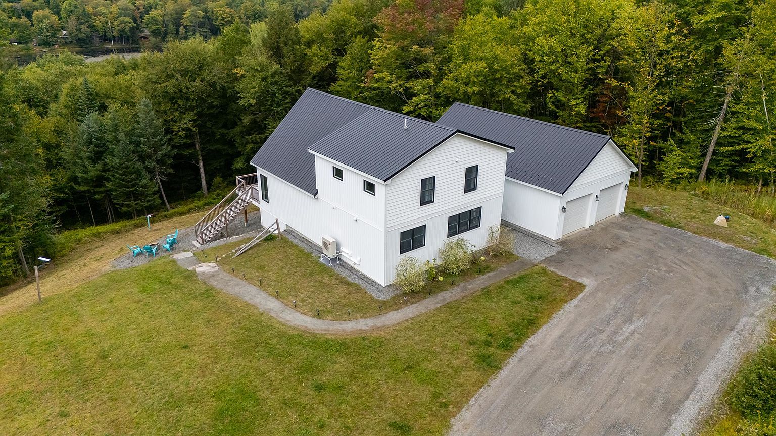 This aerial view captures a modern, white-sided home with a dark metal roof, set against a lush, wooded backdrop. The property features a gravel driveway leading to an attached two-car garage, a small deck with outdoor seating, and a sprawling green lawn. The perspective highlights the home's clean lines and its secluded, natural setting.