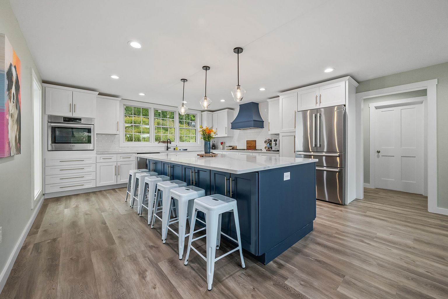 This bright and modern kitchen features a large central island with a deep blue base and a light-toned countertop, complemented by four white metal stools. The space is equipped with white cabinetry, stainless steel appliances, and a distinctive blue range hood, all set against light-colored wood-look flooring. Large windows above the sink area provide ample natural light, creating an open and inviting atmosphere.