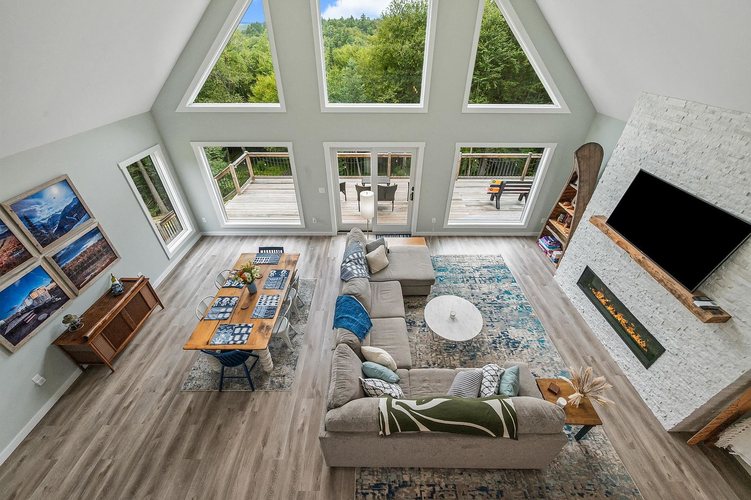 This high-angle shot captures an open-concept living and dining area characterized by a dramatic vaulted ceiling and expansive windows that frame the surrounding forest. The space features a large sectional sofa arranged on a patterned area rug, a wooden dining table, and a striking white stone-clad chimney breast housing a linear fireplace and a mounted television. The light wood-toned flooring and neutral color palette create a bright, airy, and inviting atmosphere perfect for modern mountain living.