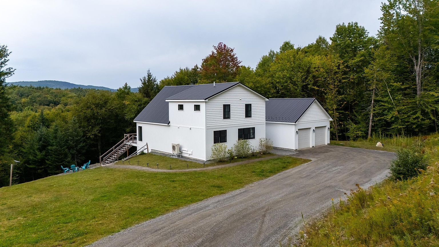 This elevated perspective captures a modern, white-sided home with a dark metal roof, nestled against a lush, forested hillside. The property features an attached two-car garage, a gravel driveway, and a grassy lawn that slopes down toward a deck area with outdoor seating. The setting is serene and rural, emphasizing the home's integration into the natural landscape.