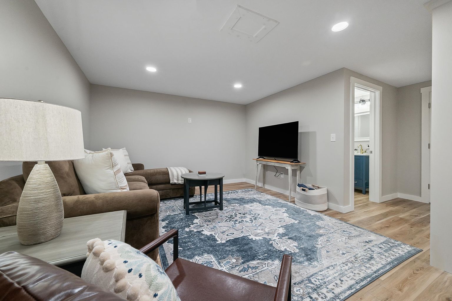 This cozy living room features light-toned wood-look flooring and a large, ornate blue and cream area rug that anchors the space. The room is furnished with a comfortable brown sectional sofa, a round coffee table, and a leather armchair, all illuminated by recessed ceiling lighting. A television sits on a slim console table, and a doorway leads into an adjacent bathroom, creating a functional and inviting layout.