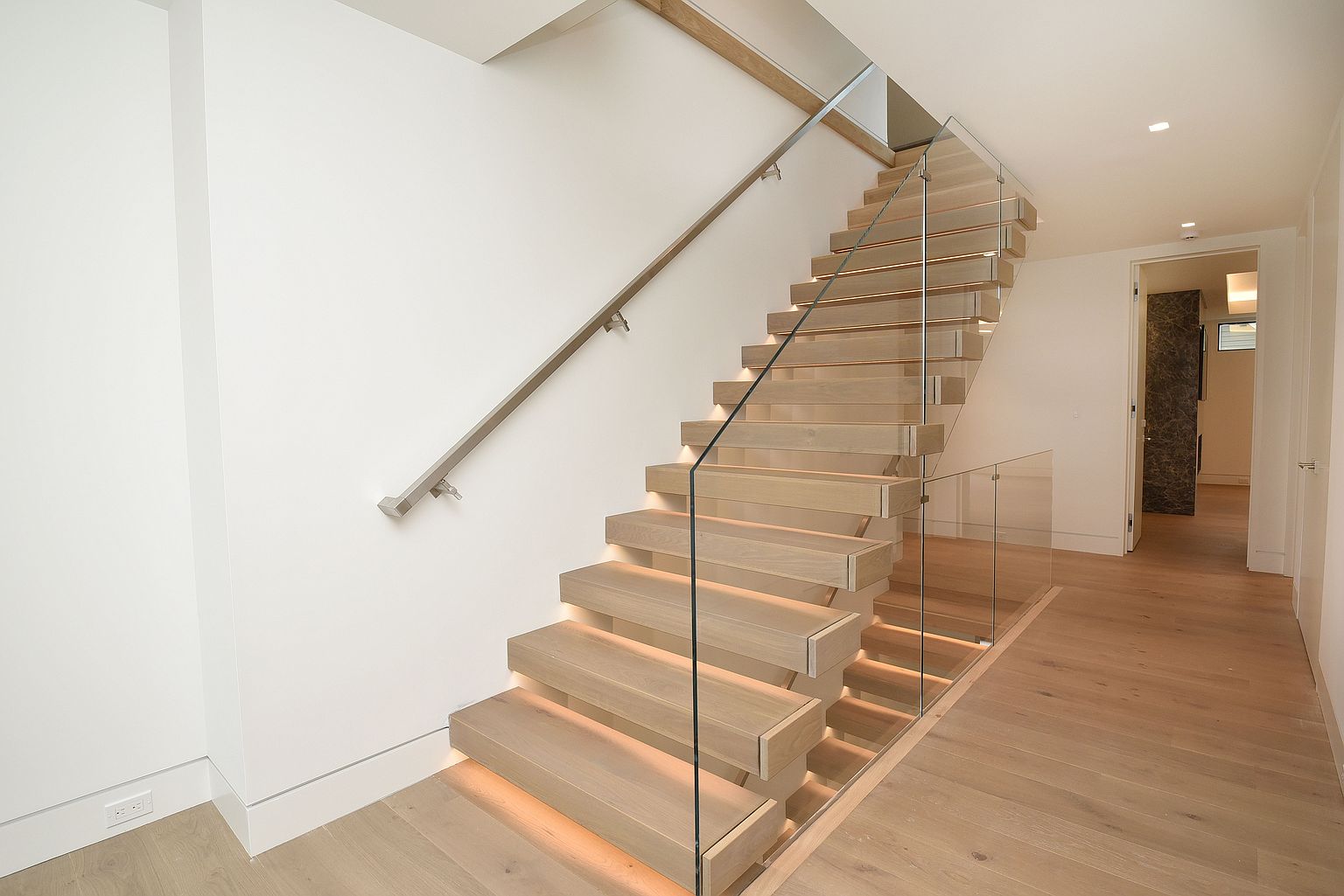 This interior shot showcases a modern staircase with floating wooden steps and glass railings, illuminated by subtle under-step lighting. The staircase ascends along a clean white wall, complemented by a sleek metal handrail. The scene leads into a bright hallway with hardwood flooring, creating a sense of contemporary elegance and open space.