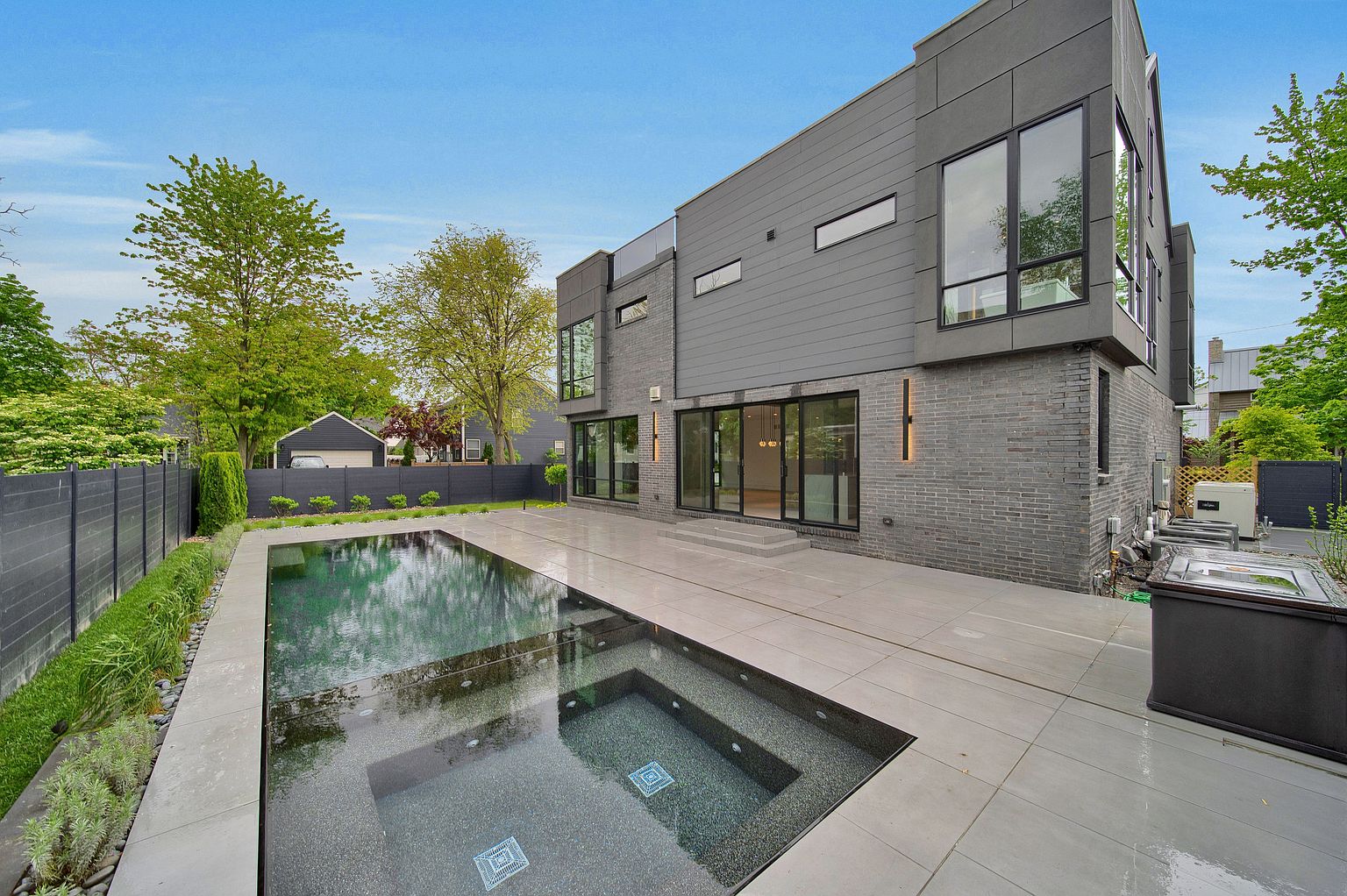 This image showcases the rear exterior of a modern home, emphasizing a sleek pool and integrated spa. The property features a contemporary design with gray brick and siding, large windows, and a spacious patio area. The pool's reflective surface and the well-maintained landscaping add to the luxurious and private backyard setting.