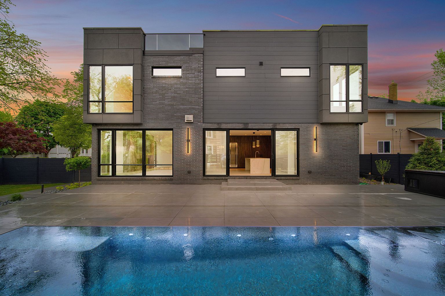 This image showcases the rear exterior of a modern home, emphasizing a luxurious pool area. The house features a gray brick and panel facade with large windows and sleek lighting fixtures. A spacious patio extends from the house to the pool, creating an inviting outdoor living space.
