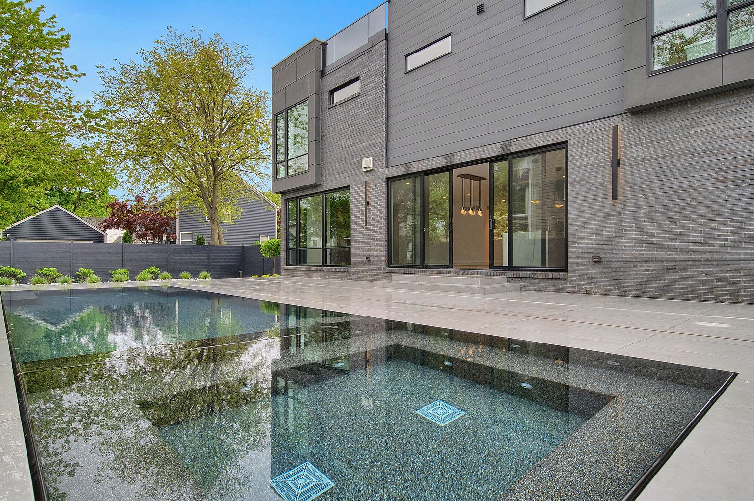 This image showcases a modern home's backyard featuring a sleek, dark-bottomed pool with a built-in spa. The pool's surface reflects the surrounding trees and the contemporary architecture of the house, which includes gray brick and siding. The patio area is paved with large, light-colored tiles, creating a clean and sophisticated outdoor living space.