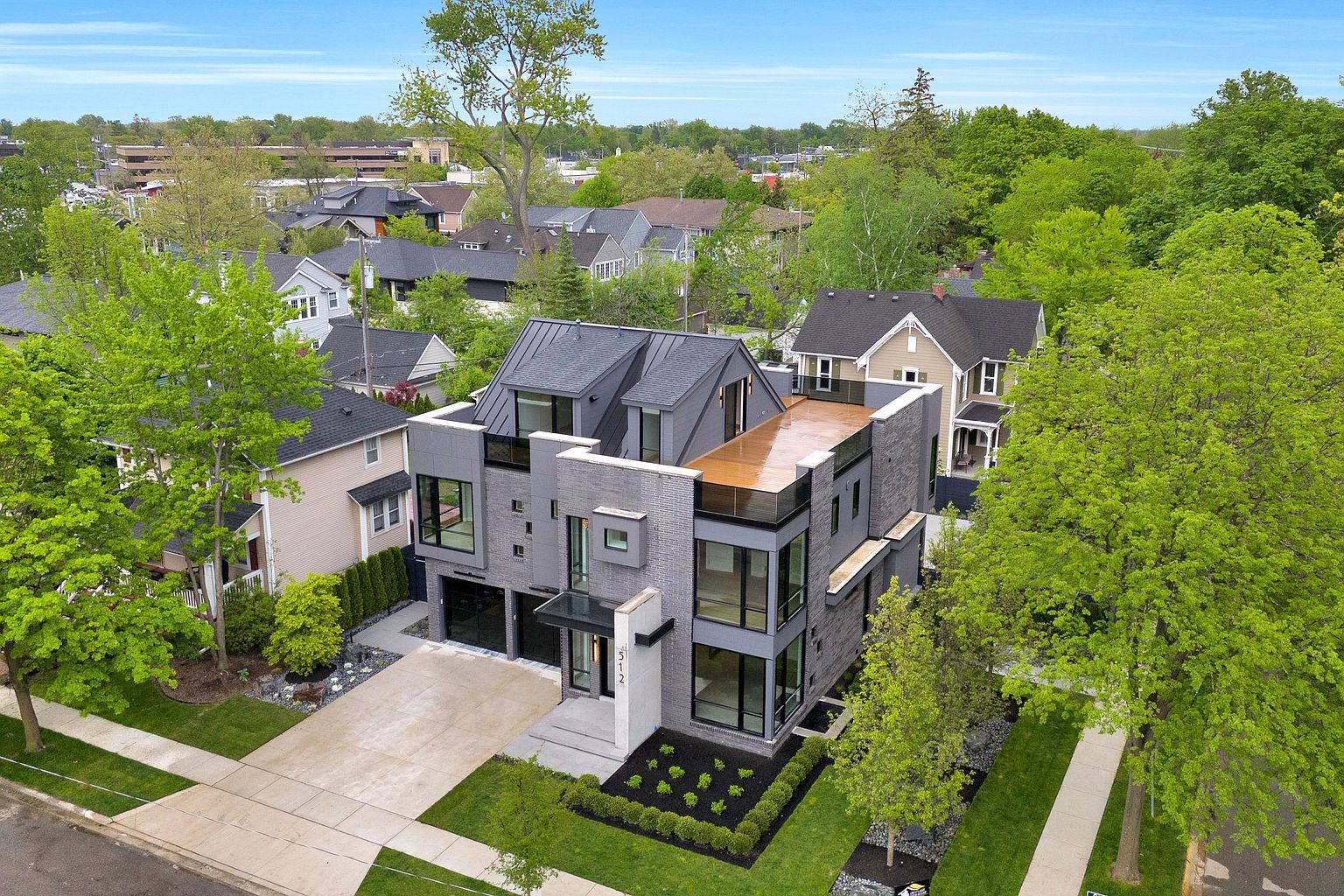This aerial view showcases a modern, multi-story home with a gray brick facade and a flat roof deck. The property features well-manicured landscaping, including a small garden bed in the front and mature trees surrounding the house. A concrete driveway leads to a two-car garage, and the surrounding neighborhood is visible in the background.