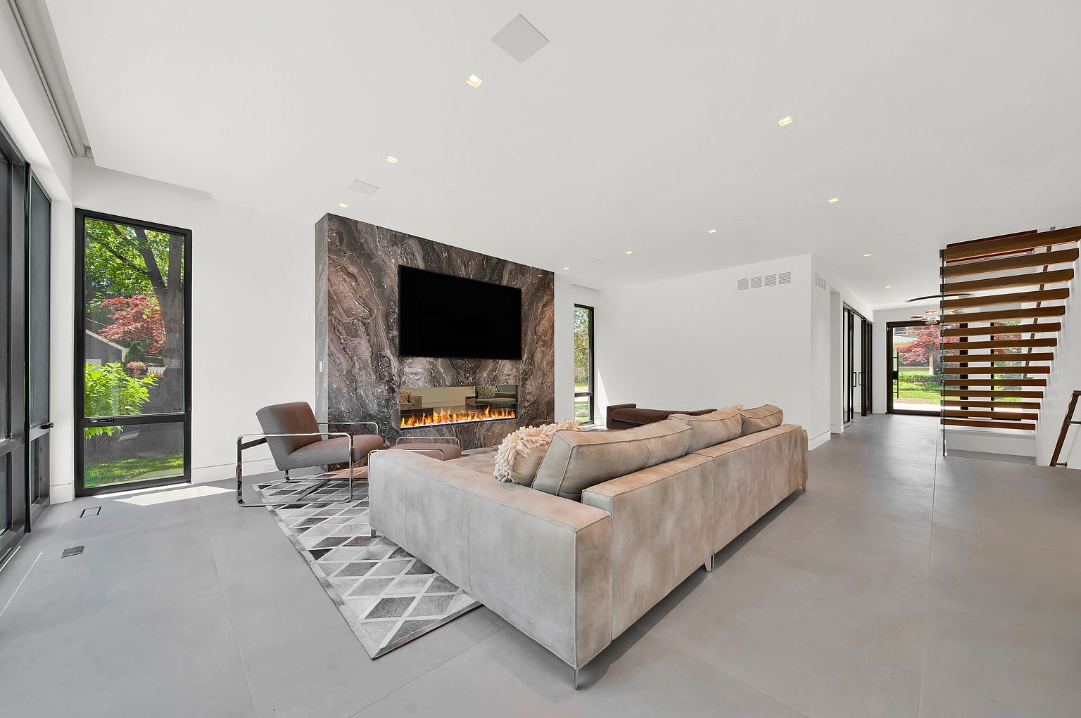 This is a modern living room featuring a large gray sofa, a marble fireplace with a built-in television, and a gray geometric rug. Large windows provide natural light and views of the outdoors. The room has a minimalist design with a focus on clean lines and neutral colors, creating a sophisticated and inviting atmosphere.