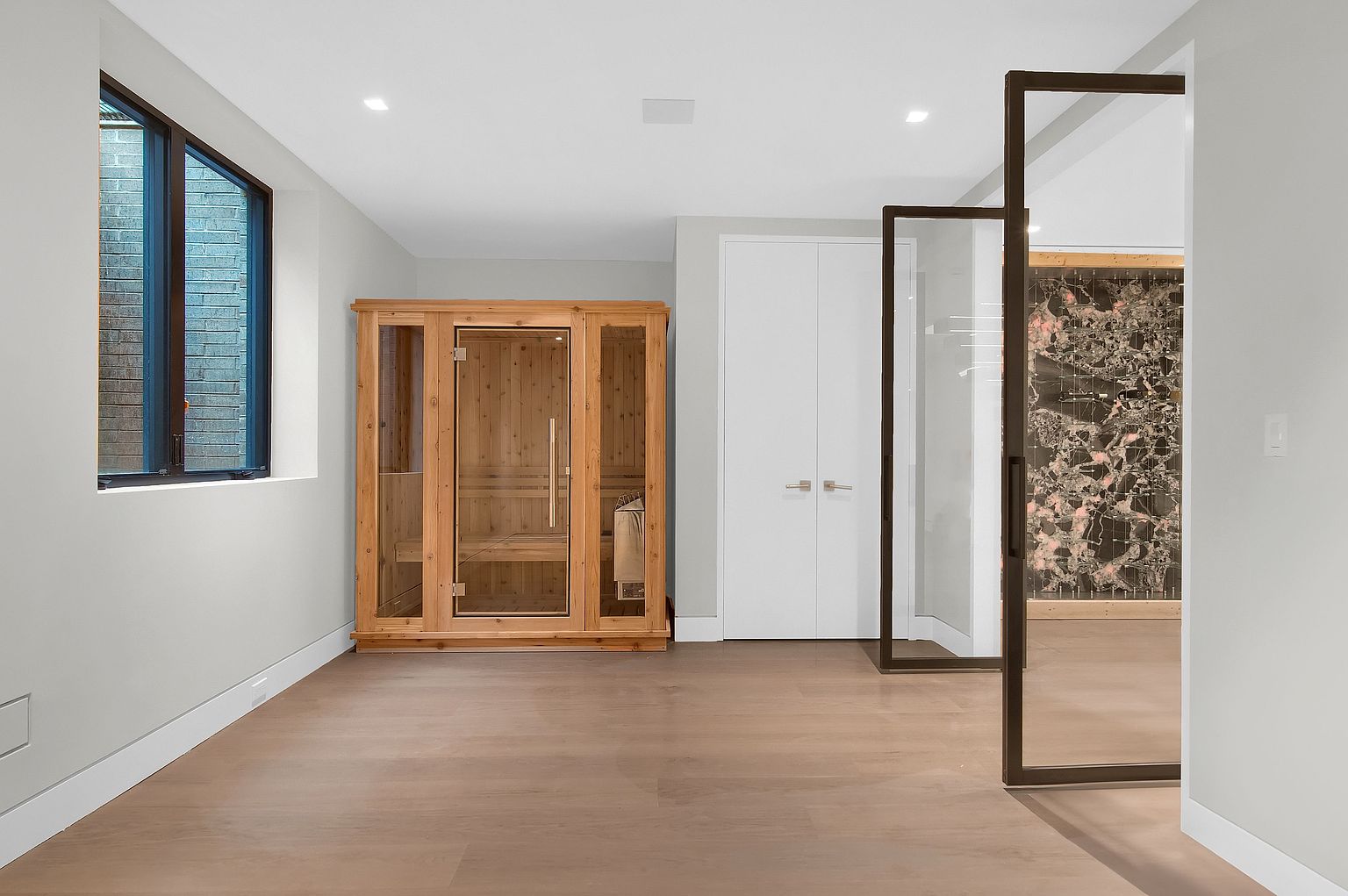 This interior shot showcases a home gym featuring a wooden sauna, light wood flooring, and a modern glass-enclosed shower area. The room is well-lit with recessed lighting and a window, creating a clean and inviting space for relaxation and wellness. The perspective is from the doorway, looking into the gym.