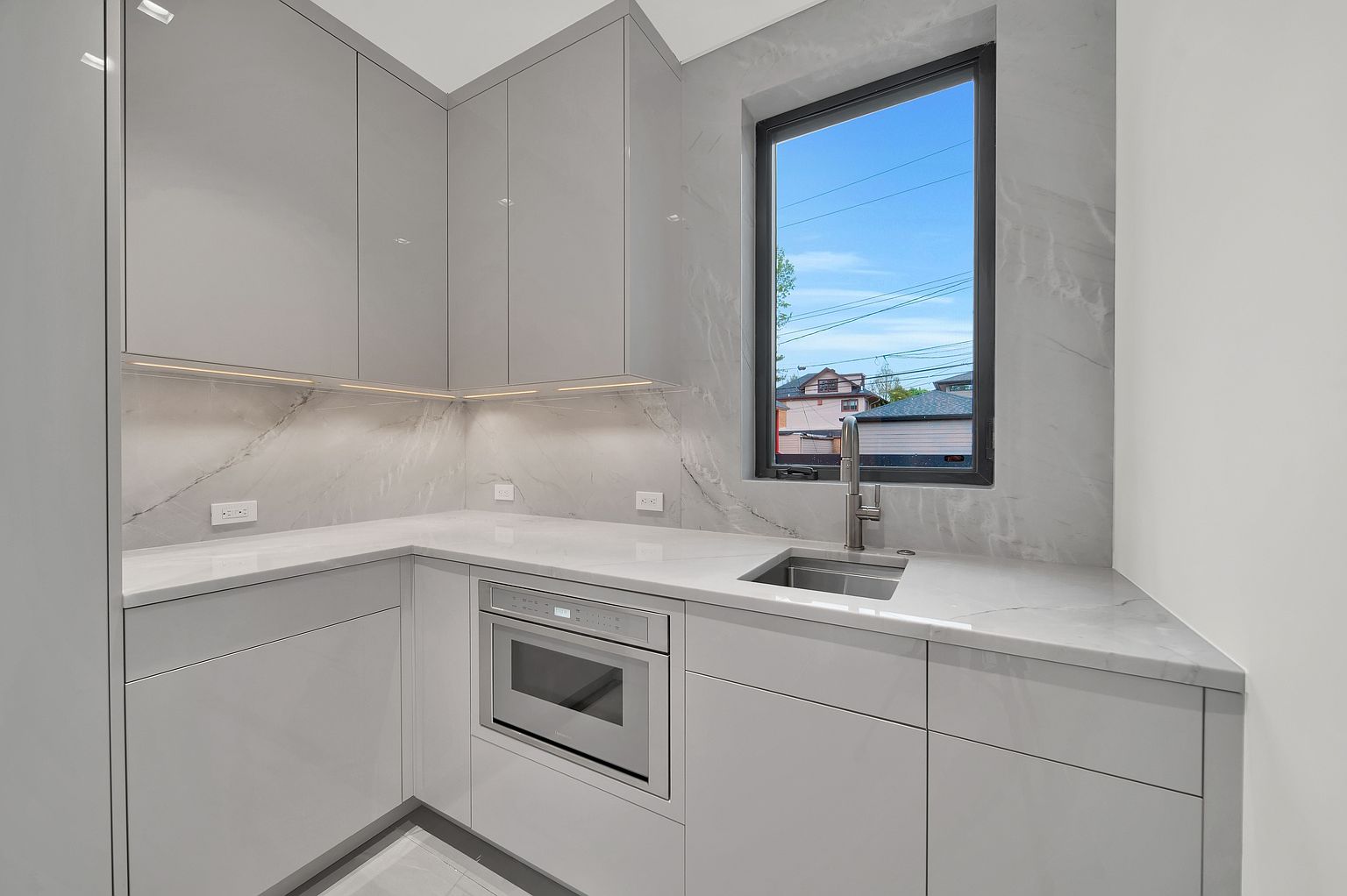 This is a modern, minimalist kitchen featuring sleek, light gray cabinetry and countertops with marble-like veining. A stainless steel sink and faucet are positioned in front of a window that offers a view of the outside. An integrated microwave is built into the cabinetry, contributing to the clean, uncluttered aesthetic.