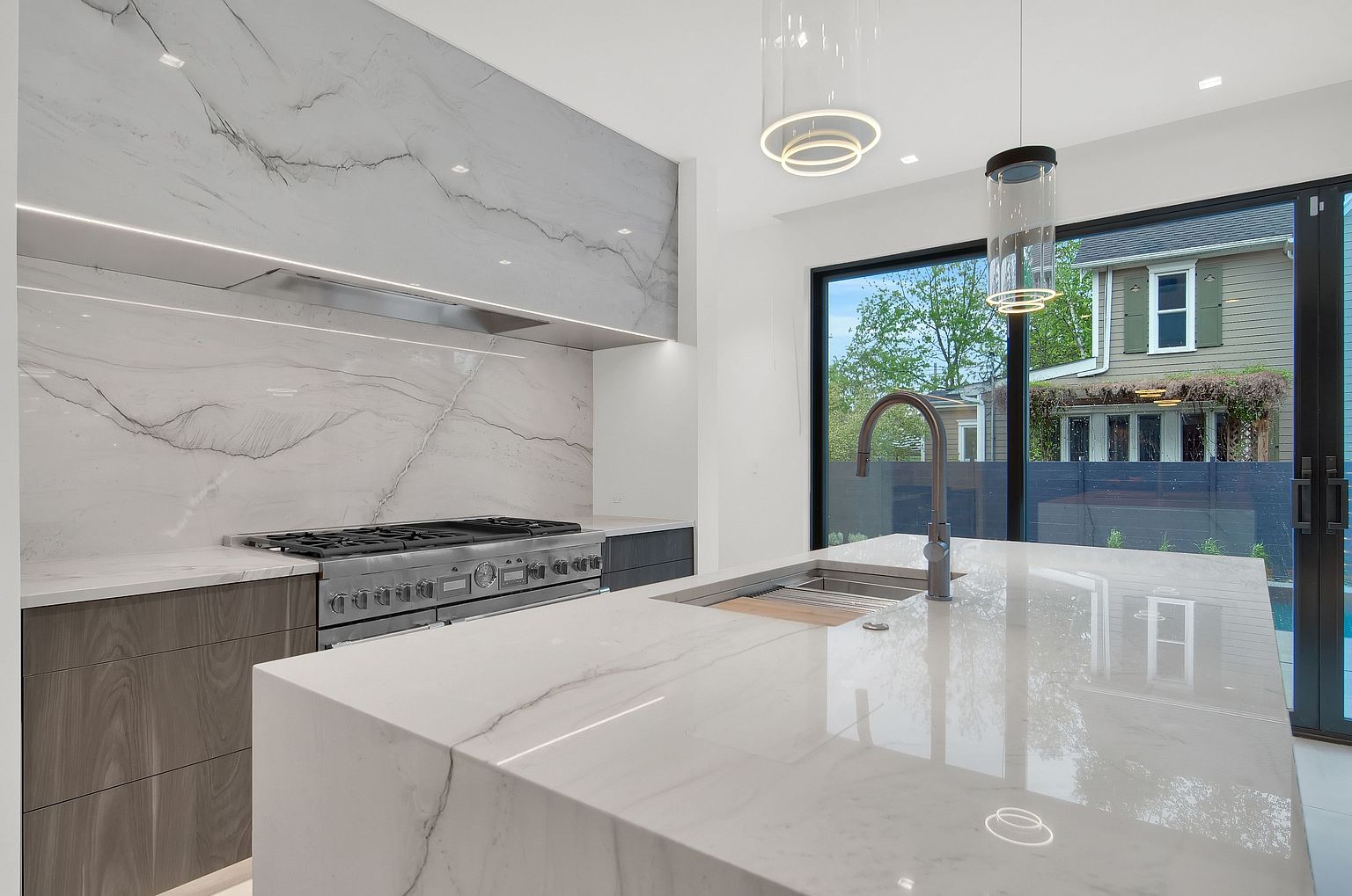 This is a modern kitchen featuring a large island with a white marble countertop and a stainless steel sink. The kitchen also features a stainless steel gas range and a marble backsplash. Large windows provide natural light and a view of the backyard.