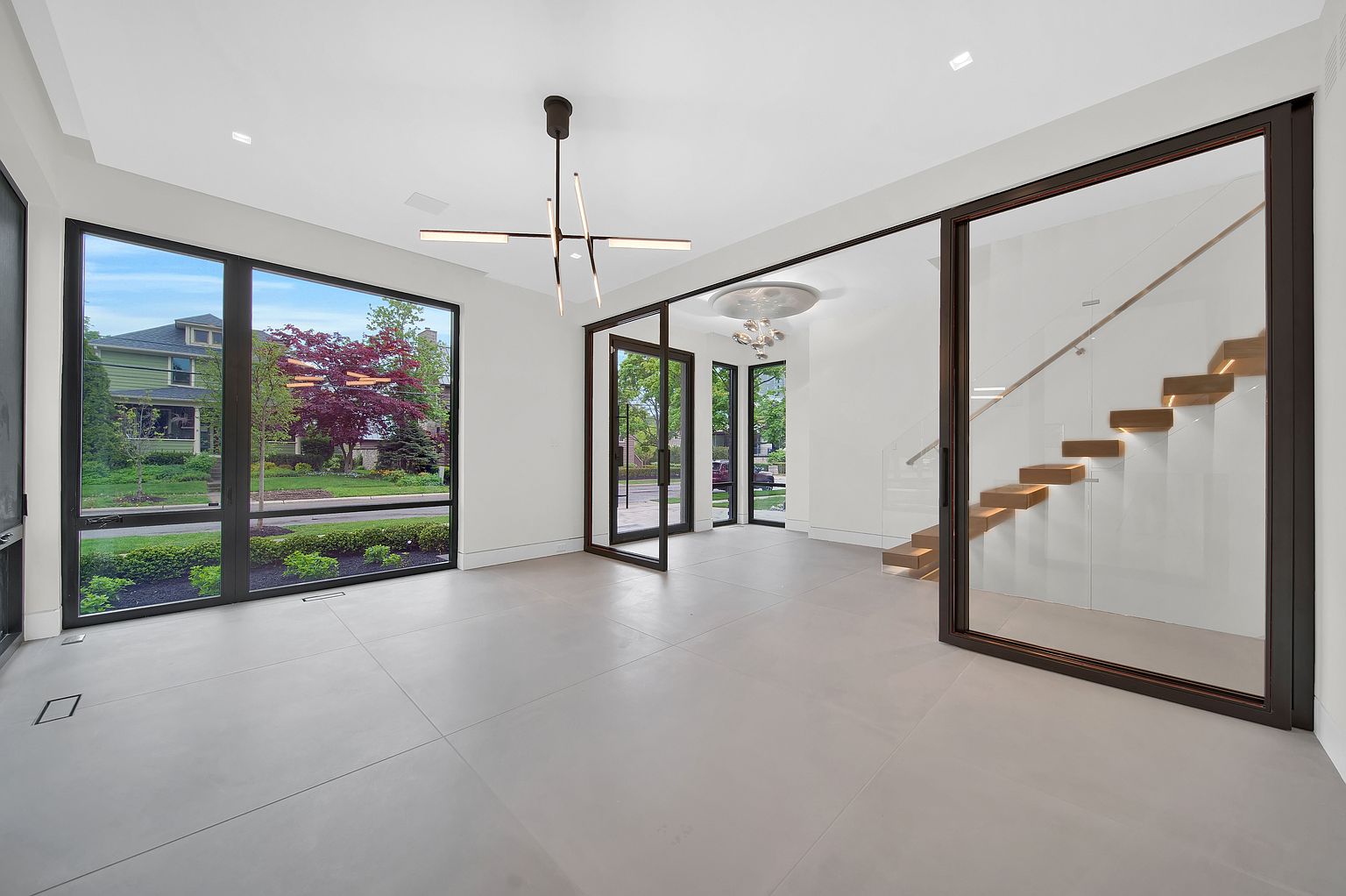 This interior shot showcases a modern hallway and staircase. The hallway features large windows offering exterior views, a contemporary chandelier, and sleek gray tile flooring. The staircase is enclosed by glass and features floating wooden steps with subtle lighting, creating a minimalist and elegant design.