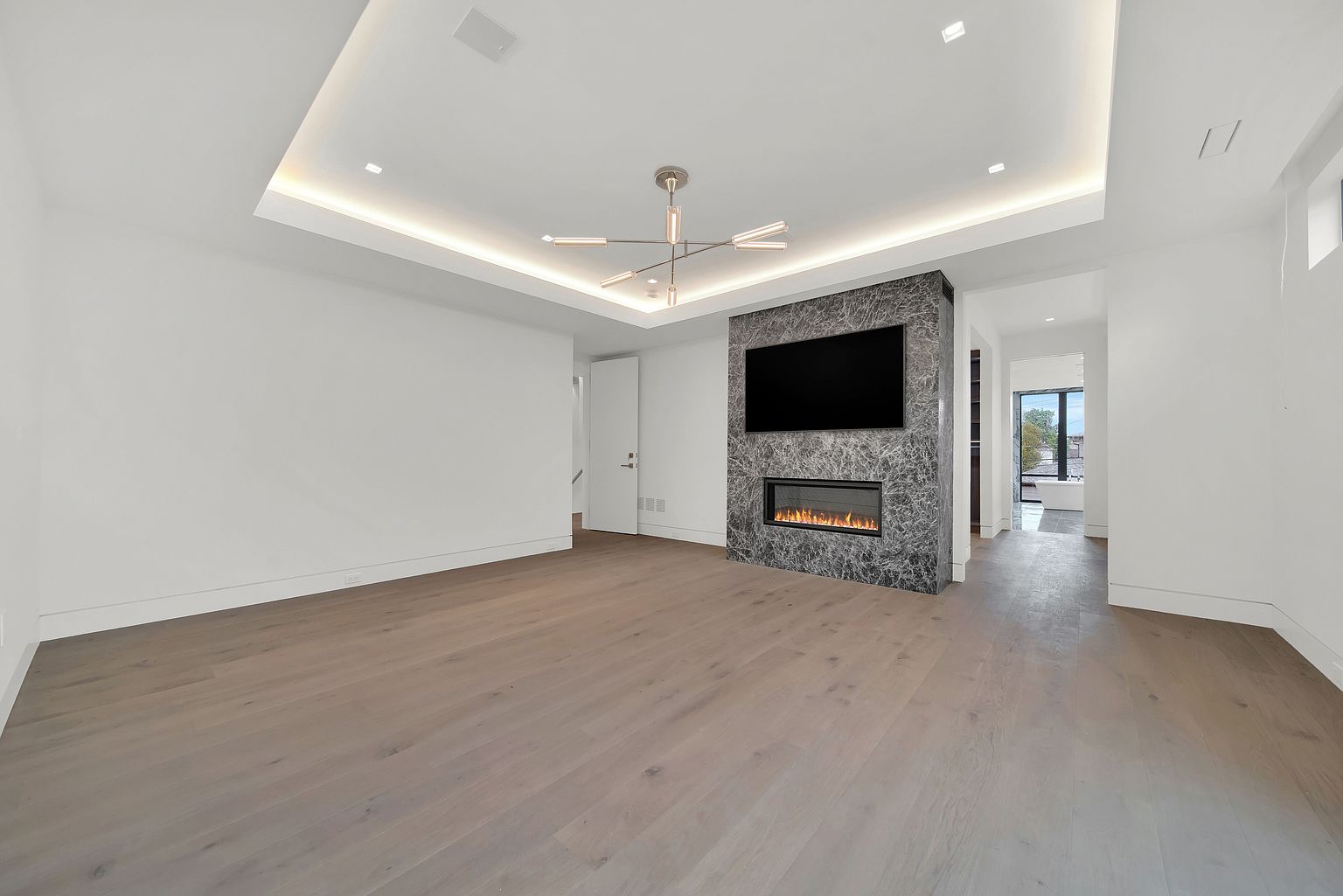 This is a spacious living room featuring light hardwood floors and white walls, creating a bright and airy atmosphere. A modern fireplace with a dark marble surround and a mounted television serves as a focal point. The room is illuminated by recessed lighting and a contemporary chandelier, enhancing the modern aesthetic.