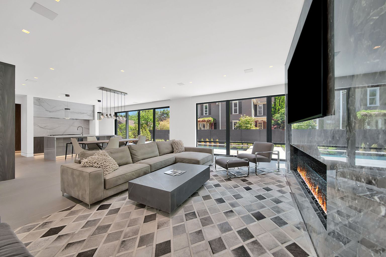 This is a spacious and modern living room featuring a large sectional sofa, a geometric patterned rug, and a sleek coffee table. Floor-to-ceiling windows provide ample natural light and views of the outdoors. A modern fireplace and TV are integrated into a marble wall, adding a luxurious touch to the space.