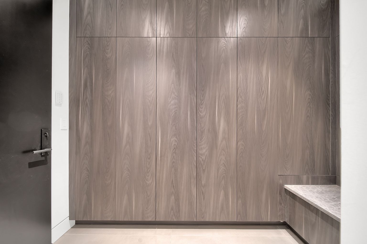 The image showcases a modern hallway featuring a sleek, dark-colored door on the left and a wall of built-in storage cabinets with a light wood grain finish on the right. A small section of countertop is visible on the right side, adding a touch of functionality. The overall aesthetic is clean, minimalist, and contemporary, suggesting a well-designed and organized space.