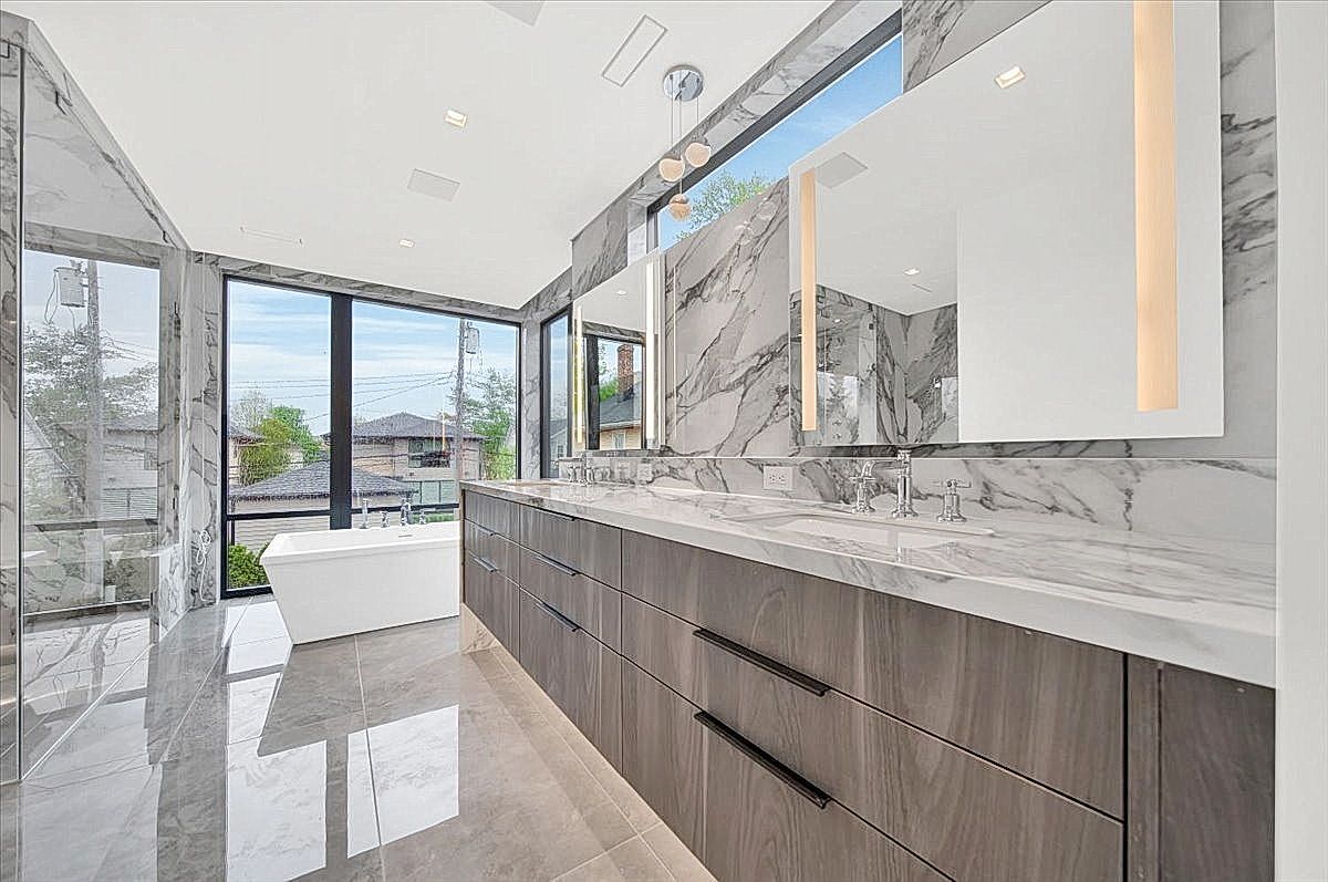 This is a luxurious primary bathroom featuring a long vanity with gray wood-grain cabinets and a marble countertop. Two large mirrors with integrated lighting are mounted above the sinks, and the walls are covered in marble. A freestanding bathtub is visible near a large window, and the floor is tiled with gray stone, creating a modern and elegant space.