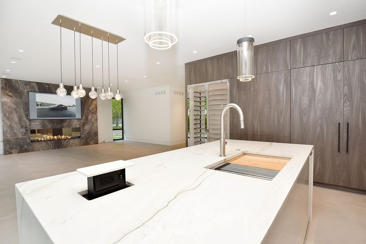 This is a modern kitchen featuring a large island with a white countertop and a stainless steel sink. The kitchen also has wooden cabinets and a wine cooler. The room is well-lit with pendant lights and recessed lighting, and the overall style is sleek and contemporary.