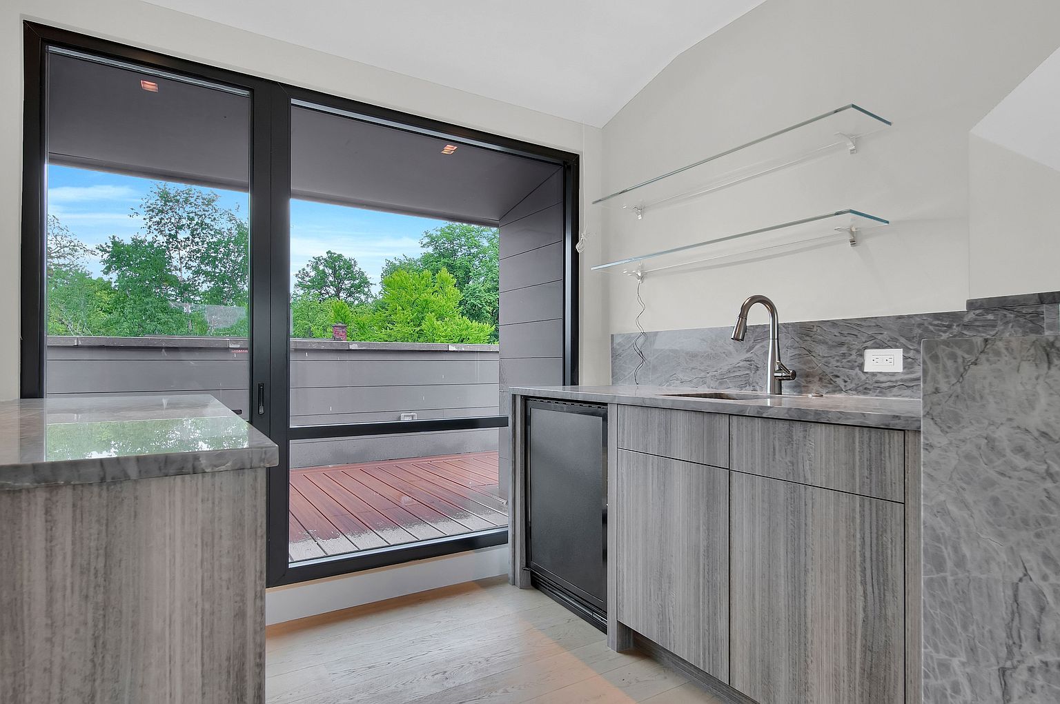 This interior shot showcases a modern kitchen area with sleek gray cabinetry and countertops, complemented by stainless steel fixtures. A large sliding glass door provides access to an outdoor deck, offering a seamless indoor-outdoor transition and views of lush greenery. The space is well-lit and appears to be designed for both functionality and aesthetic appeal.