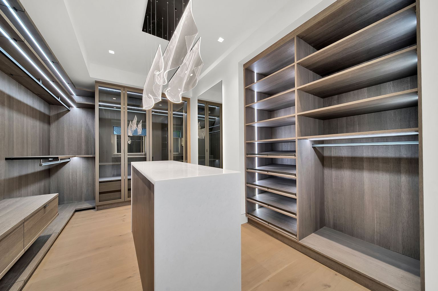 This is a well-organized walk-in closet featuring custom shelving and drawers in a modern style. The closet includes a central island with a white countertop, providing ample space for folding and organizing clothes. Integrated lighting enhances the luxurious feel, and the wood flooring adds warmth to the space.