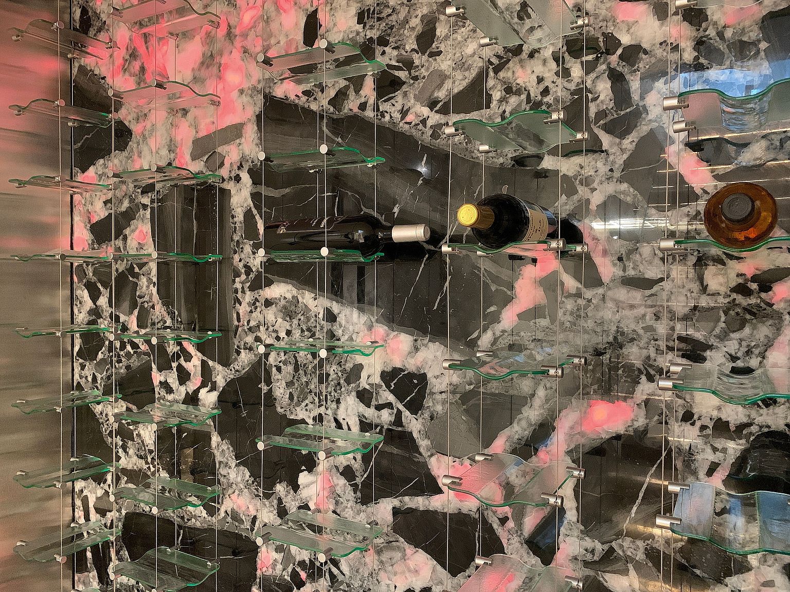 This image showcases a modern wine cellar featuring a striking marble wall as a backdrop. Wine bottles are displayed on sleek, transparent glass shelves supported by a minimalist metal framework. The lighting casts a soft glow, highlighting the textures and colors of the marble and the wine bottles, creating an elegant and sophisticated ambiance.