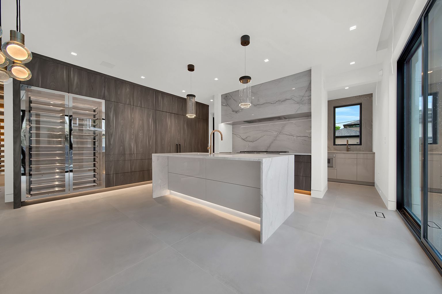 This is a modern kitchen with a large island featuring a waterfall countertop and under-cabinet lighting. The kitchen includes dark wood cabinetry, a wine fridge, and pendant lighting above the island. The space is open and bright, with large format tile flooring and a sliding glass door leading to the outside.