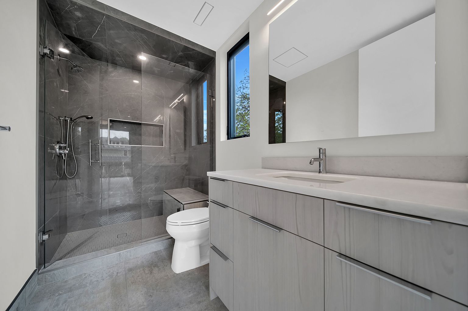 This is a modern primary bathroom featuring a glass-enclosed shower with gray marble-like tiling and a built-in bench. A sleek vanity with light wood-tone cabinets and a white countertop is complemented by a large, illuminated mirror. The overall design is minimalist and luxurious, creating a spa-like atmosphere.