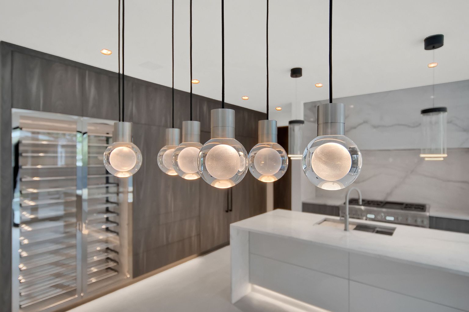 This is a modern kitchen featuring a white island with a marble countertop and stainless steel appliances. The kitchen is illuminated by several pendant lights with spherical glass shades. A wine fridge is built into the wall, and the overall design is sleek and contemporary.