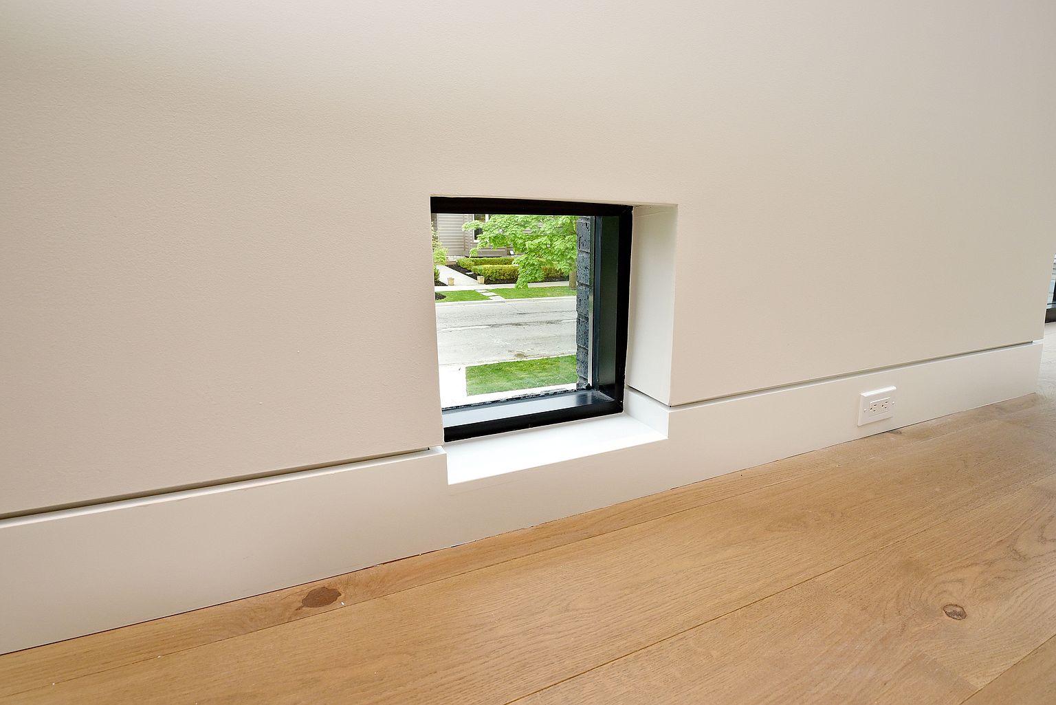 This interior shot showcases a unique architectural detail: a small, square window set low in the wall, offering a glimpse of the exterior. The room features a clean, minimalist design with white walls, light wood flooring, and a modern baseboard. An electrical outlet is visible on the wall, emphasizing the functional aspect of the space.