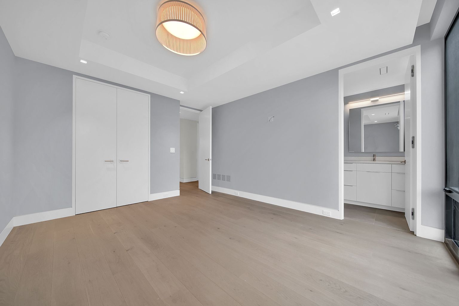 This is a spacious primary bedroom featuring light hardwood flooring and light gray walls. A large double-door closet is present, along with an open doorway leading to another room and an entrance to a bathroom. A modern, circular light fixture illuminates the room, creating a serene and inviting atmosphere.