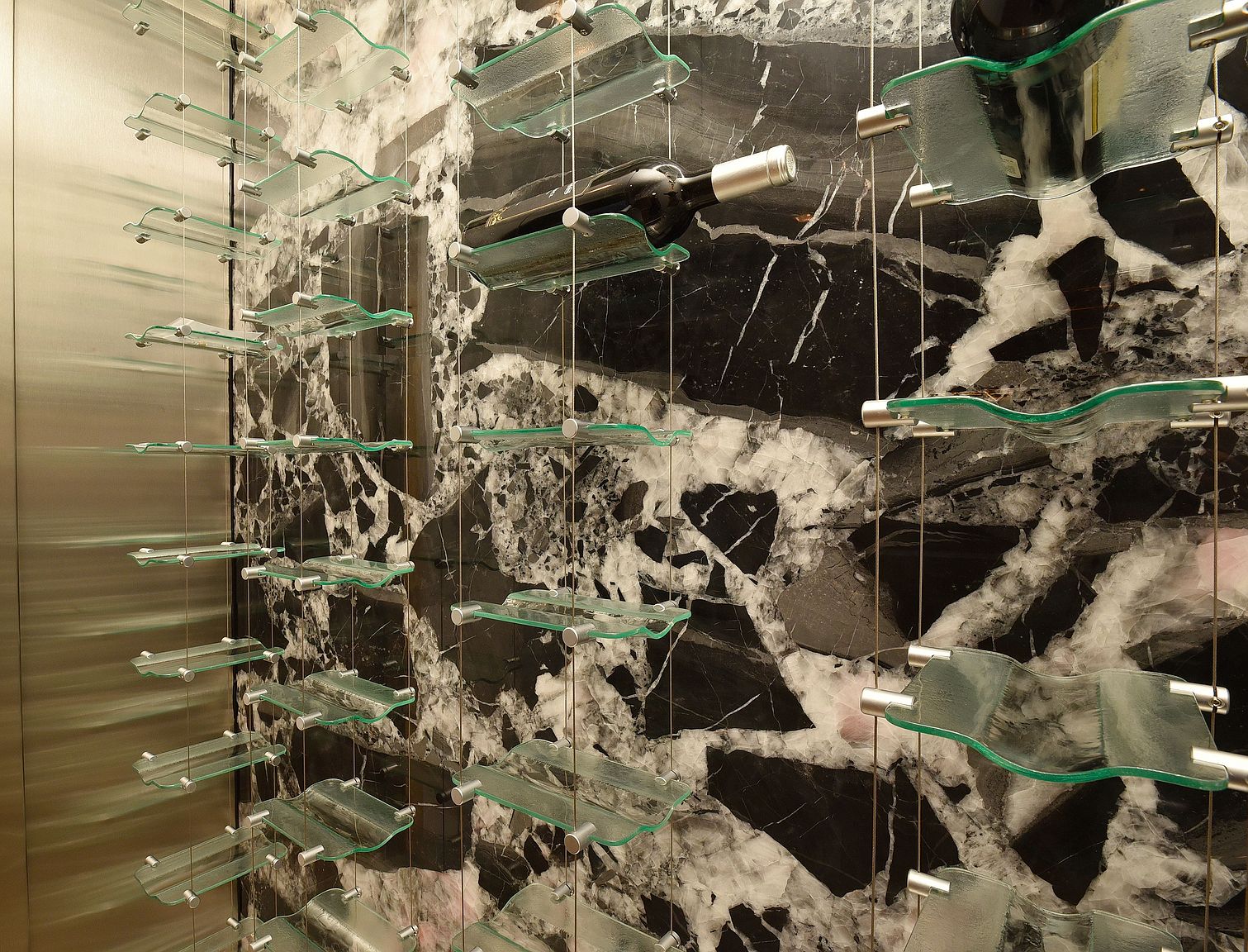 This image showcases a modern wine cellar featuring a unique display system with glass shelves suspended by metal cables against a striking marble wall. A wine bottle is visible on one of the shelves, highlighting the cellar's functionality. The overall design is sleek and sophisticated, creating an elegant storage solution for wine enthusiasts.