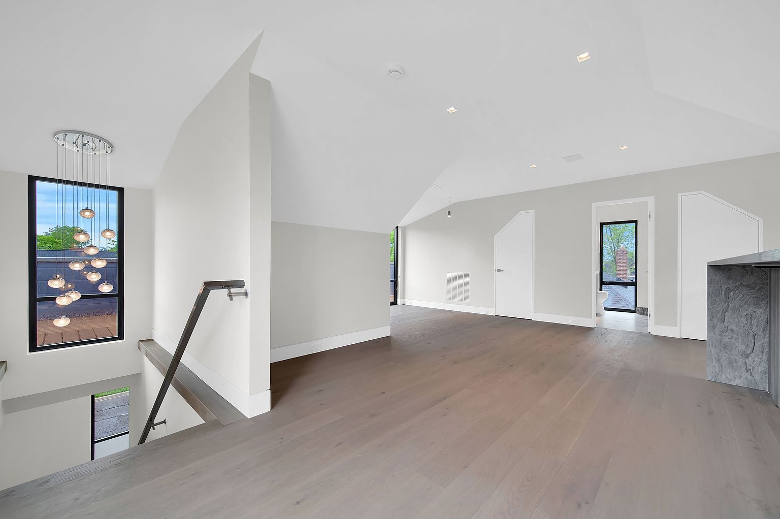 This interior shot showcases a modern hallway and staircase area. The space features light gray hardwood flooring, white walls, and a contemporary chandelier hanging above the staircase. A large window provides natural light, and the overall design is clean and minimalist.