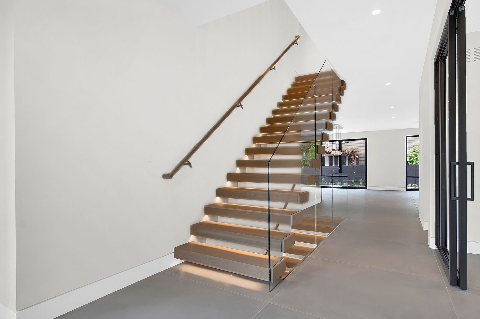 This interior shot showcases a modern hallway with a striking floating staircase. The staircase features wooden treads, glass railings, and subtle under-step lighting, creating a sleek and contemporary design. The hallway is bright and spacious, with light gray flooring and a minimalist aesthetic, leading to a view of the outdoors through large windows.