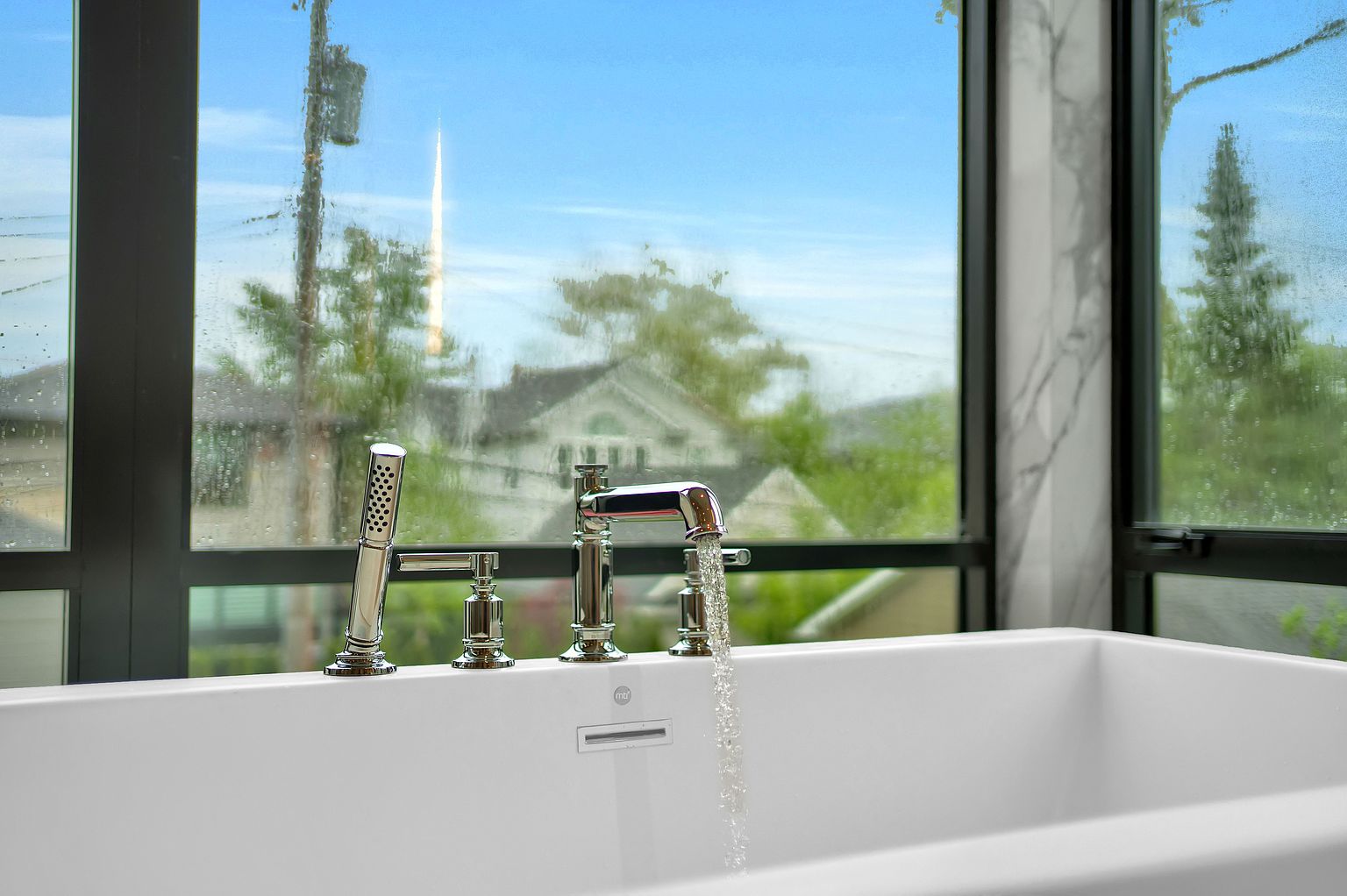 This image showcases a luxurious primary bathroom featuring a modern freestanding bathtub with a sleek chrome faucet and handheld shower. Large windows offer a view of the outdoors, creating a serene and spa-like atmosphere. The overall impression is one of elegance and relaxation.