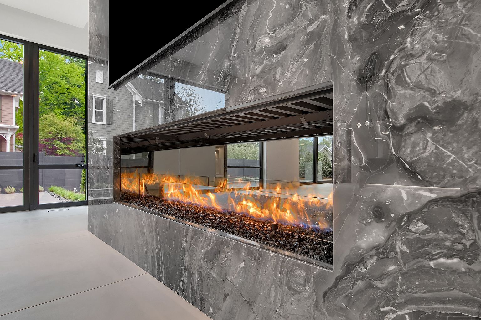 This interior shot showcases a modern living room with a striking marble fireplace. The fireplace features a long, horizontal firebox filled with black stones and vibrant flames, creating a warm and inviting ambiance. Large windows provide natural light and a glimpse of the exterior, while a sleek, minimalist design aesthetic prevails throughout the space.