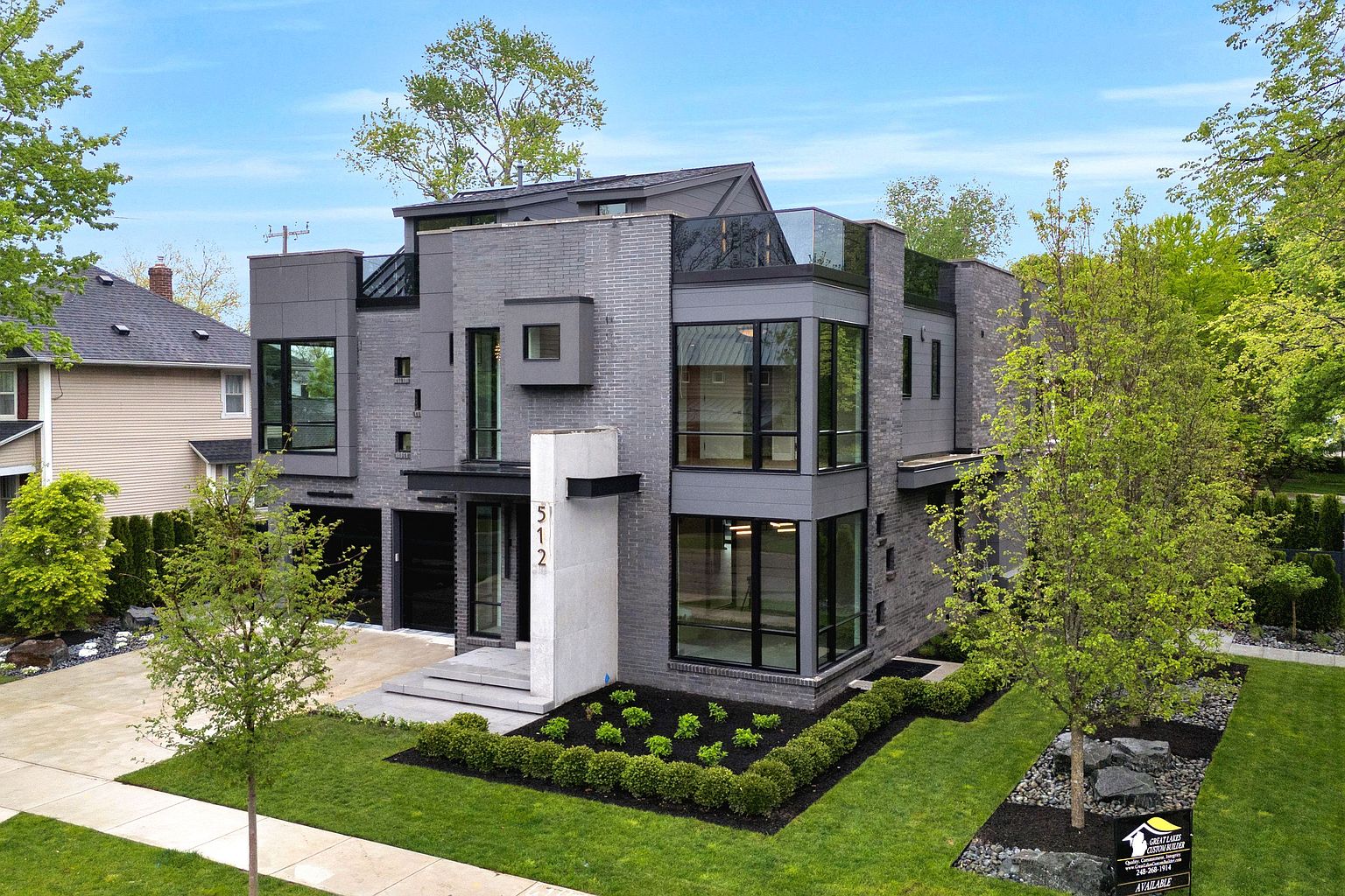 This is a front exterior view of a modern, multi-story home. The house features a gray brick and panel facade with large, dark-framed windows. The landscaping includes a well-manicured lawn, trimmed bushes, and young trees, enhancing the property's curb appeal.