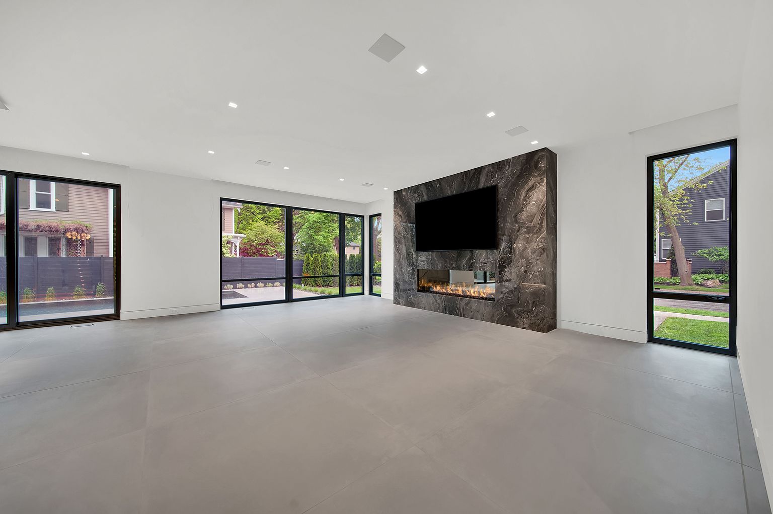 This is a spacious and modern living room featuring large windows that provide ample natural light and views of the outdoors. A striking dark marble fireplace with a built-in television serves as a focal point, adding a touch of luxury. The room is characterized by its minimalist design, clean lines, and neutral color palette, creating a sophisticated and inviting atmosphere.
