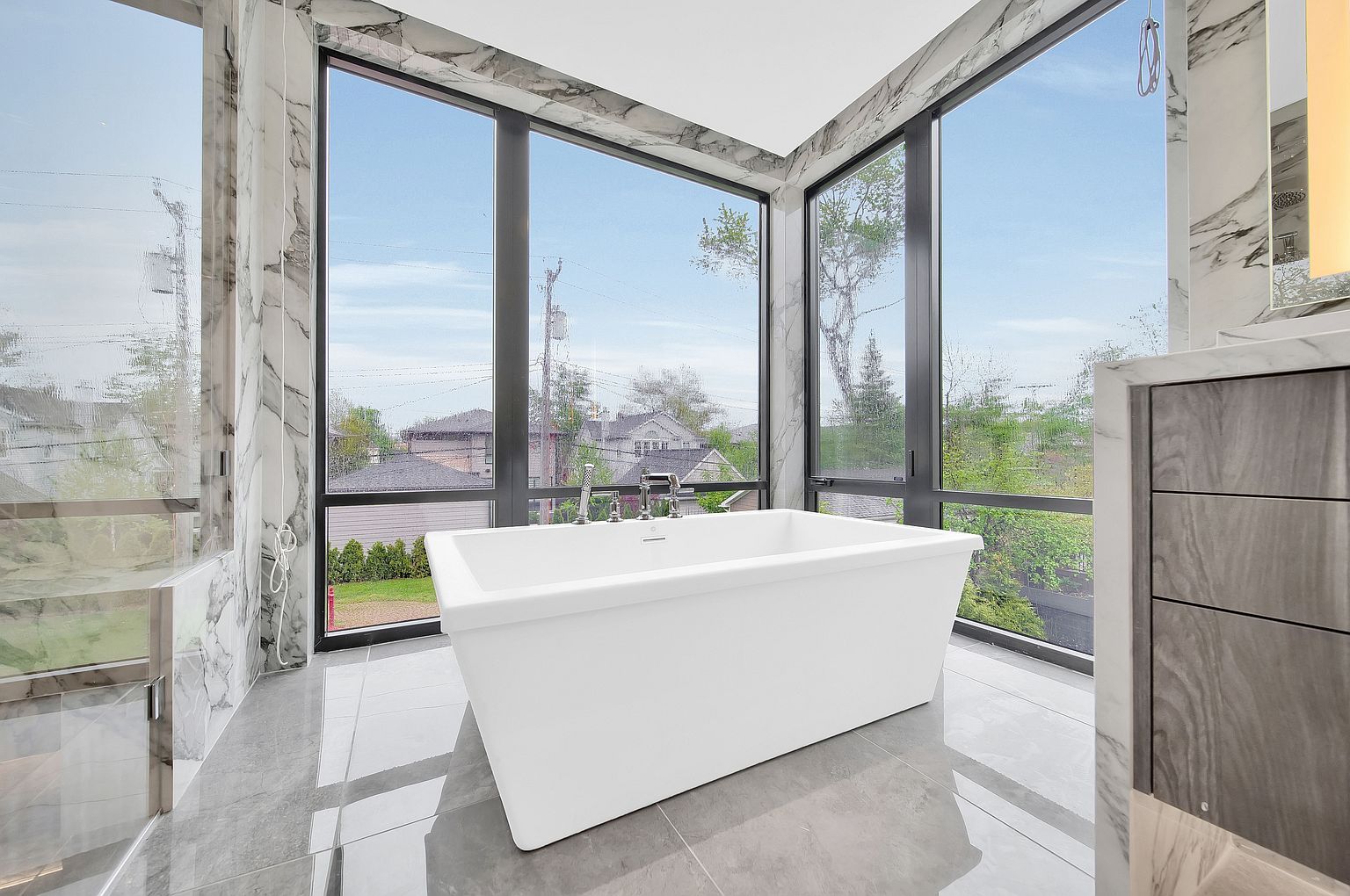 This is a luxurious primary bathroom featuring a freestanding white bathtub positioned in front of large windows offering a scenic view. The bathroom is adorned with marble accents and gray tile flooring, creating a modern and elegant aesthetic. A vanity with gray wood drawers is visible on the right side of the image.