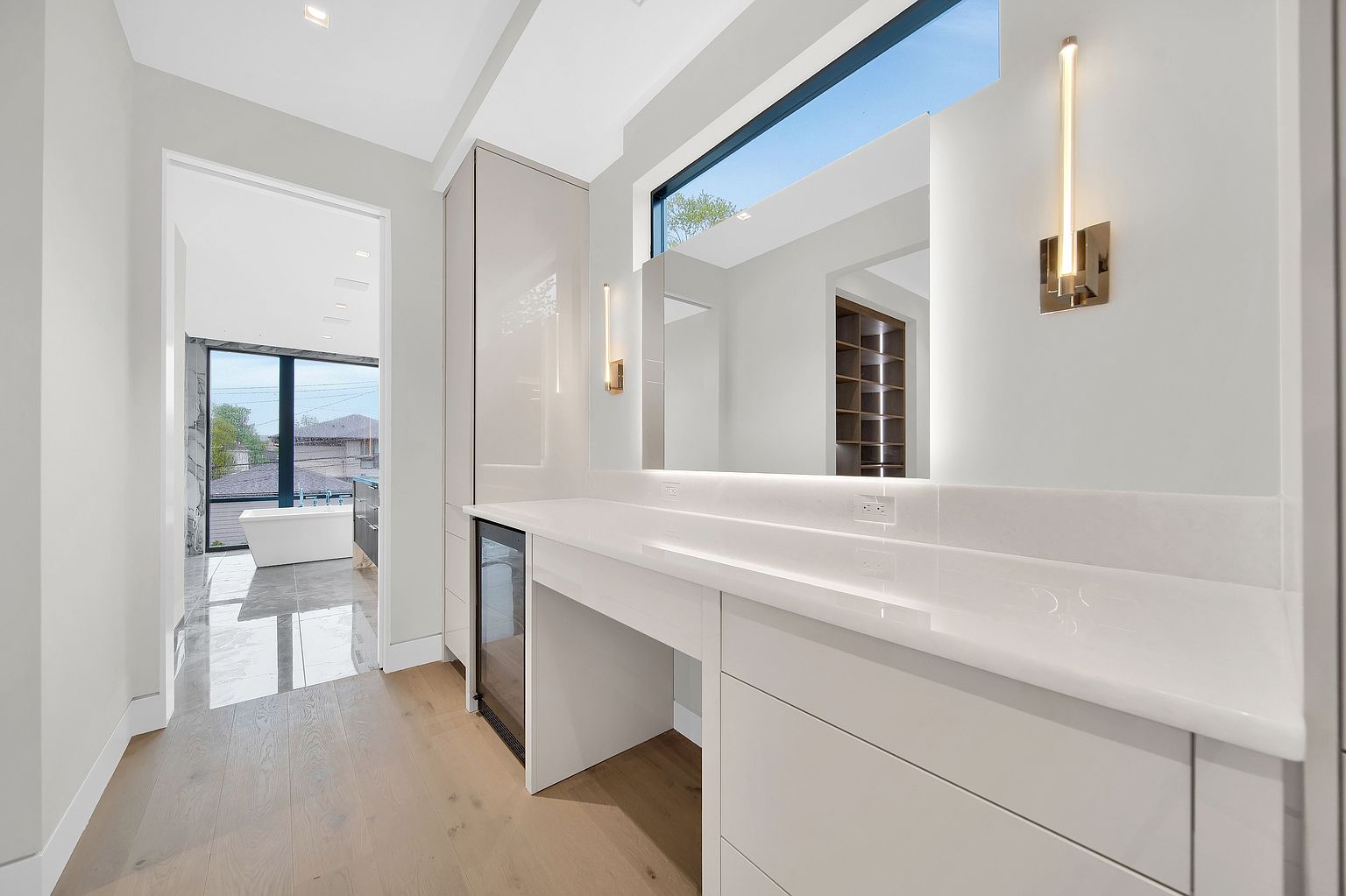 This is a bright and modern primary bathroom featuring a sleek vanity with white countertops and cabinetry. A large mirror reflects natural light from a window above, and stylish sconces flank the mirror. The bathroom leads into another area with a freestanding tub, creating a spa-like atmosphere.
