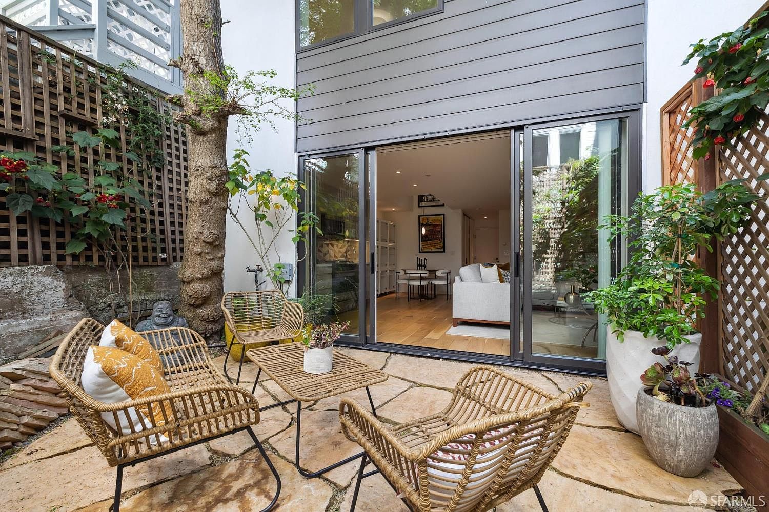 This inviting outdoor patio features natural stone pavers and a set of modern, woven rattan-style furniture, creating a cozy lounge area. The space is framed by wooden lattice trellises adorned with climbing greenery and a mature tree, offering a private, garden-like atmosphere. Large sliding glass doors provide a seamless transition into the bright, open-concept interior living space, highlighting a strong indoor-outdoor connection.