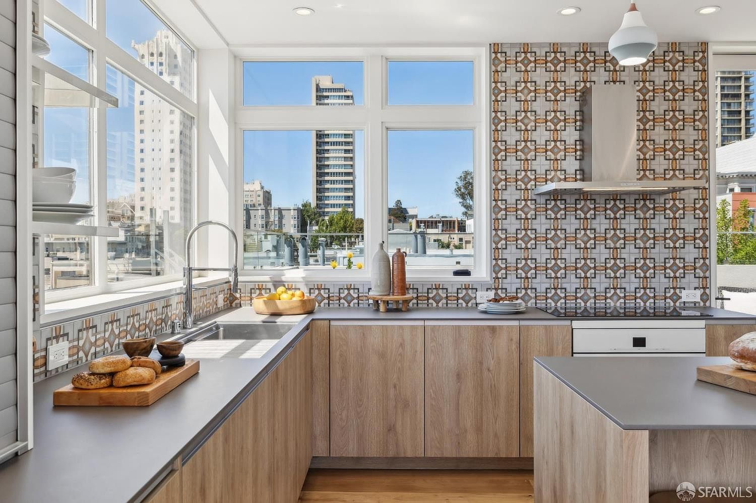 This modern kitchen features light wood-toned cabinetry paired with dark, solid-surface countertops and a striking patterned tile backsplash that extends behind the range. Large windows frame an urban cityscape, flooding the space with natural light and providing an open, airy feel. The layout includes a functional corner sink area and a central island, creating a clean and contemporary aesthetic.