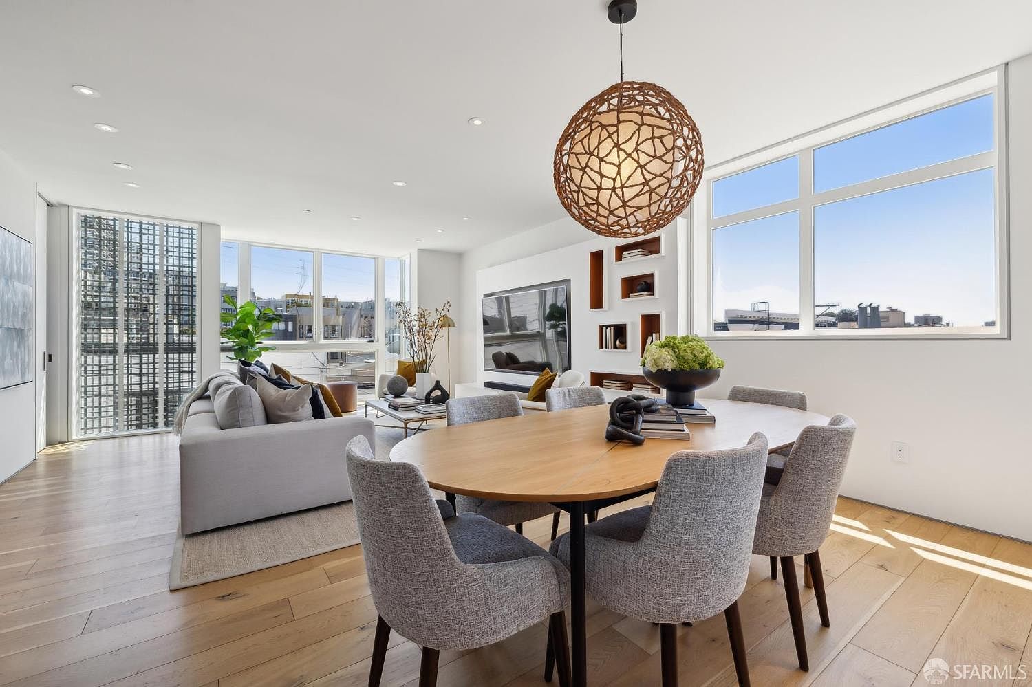 This open-concept living and dining area features light wood flooring and a bright, airy atmosphere enhanced by large windows that provide ample natural light. The space is anchored by a contemporary oval dining table with grey upholstered chairs and a cozy living area with a plush sofa and a textured, spherical pendant light. A wall-mounted television is integrated into a custom shelving unit, creating a sleek and modern aesthetic suitable for sophisticated urban living.