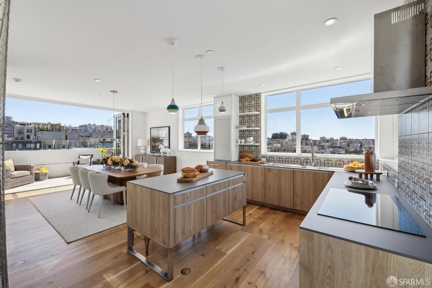 This bright, open-concept kitchen and dining area features light wood cabinetry, a central island with a dark countertop, and a sleek induction cooktop. Large windows and a folding glass door system provide an expansive view of the city skyline and access to a private balcony. The space is accented by patterned tile backsplashes and modern pendant lighting, creating a sophisticated and airy atmosphere.