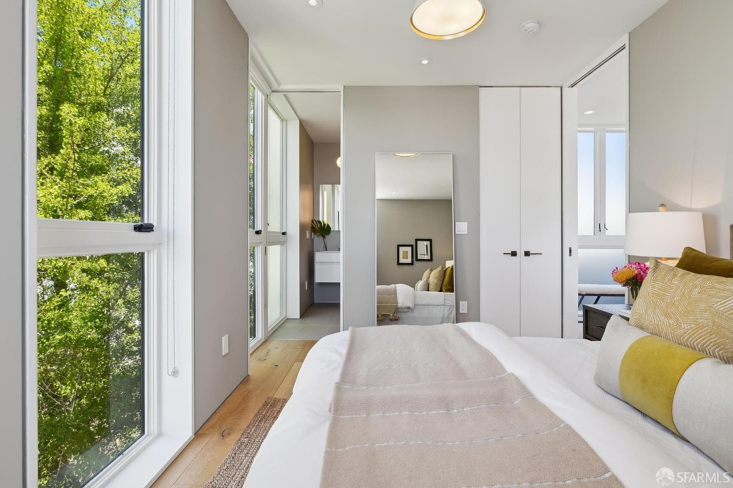 This bright and modern bedroom features light wood flooring and a neutral color palette, with a large window providing a view of lush green trees outside. The room includes a plush bed with patterned pillows, a tall mirror reflecting the adjacent bathroom, and a white closet unit. The space feels airy and serene, with a contemporary design that emphasizes natural light and clean lines.