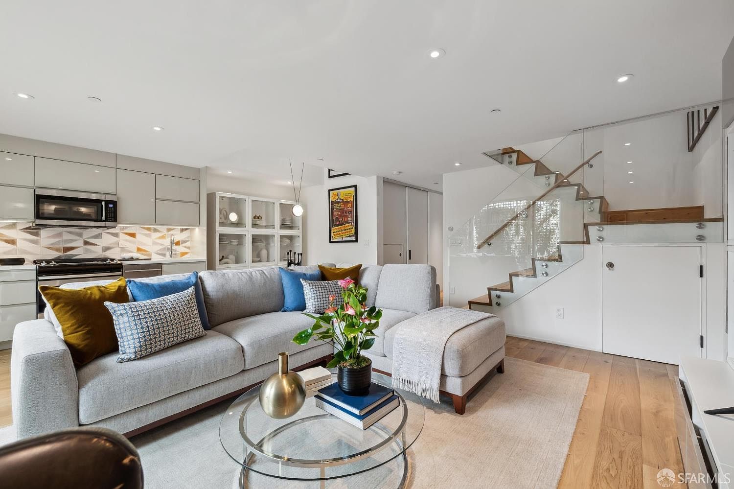 This open-concept living space features a light gray sectional sofa accented with colorful throw pillows, centered around a glass-topped coffee table. To the left, a modern kitchen showcases sleek cabinetry and a geometric patterned backsplash, while a staircase with glass railings and light wood treads ascends to the right. The room is bright and airy, with recessed lighting and light-toned wood flooring creating a cohesive, contemporary aesthetic.