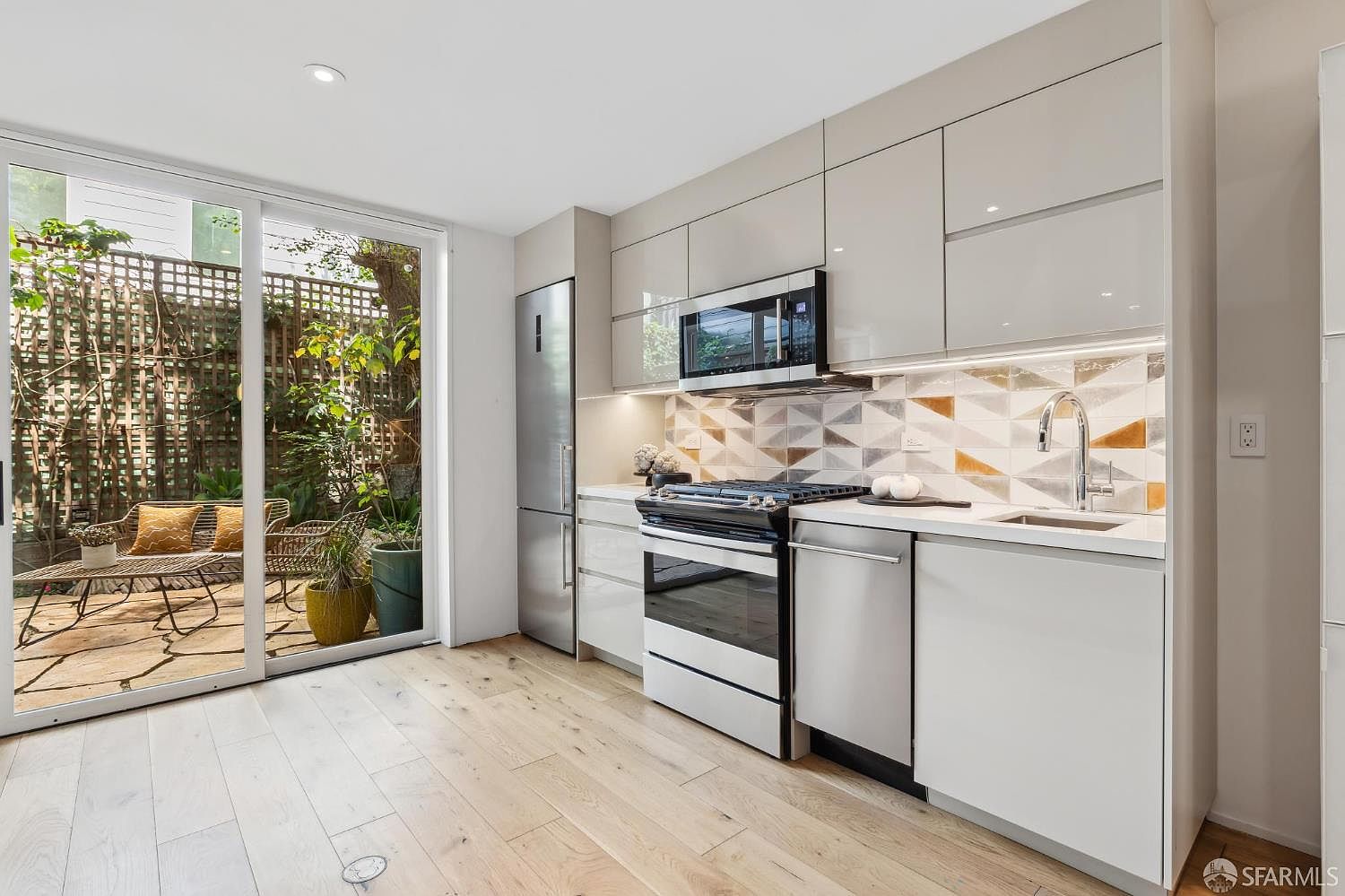 This modern, compact kitchen features sleek, light-toned cabinetry, stainless steel appliances, and a geometric patterned tile backsplash. The space is brightened by natural light streaming through large glass sliding doors that provide a seamless transition to an outdoor patio area. The light wood flooring and clean lines create a contemporary and airy atmosphere.