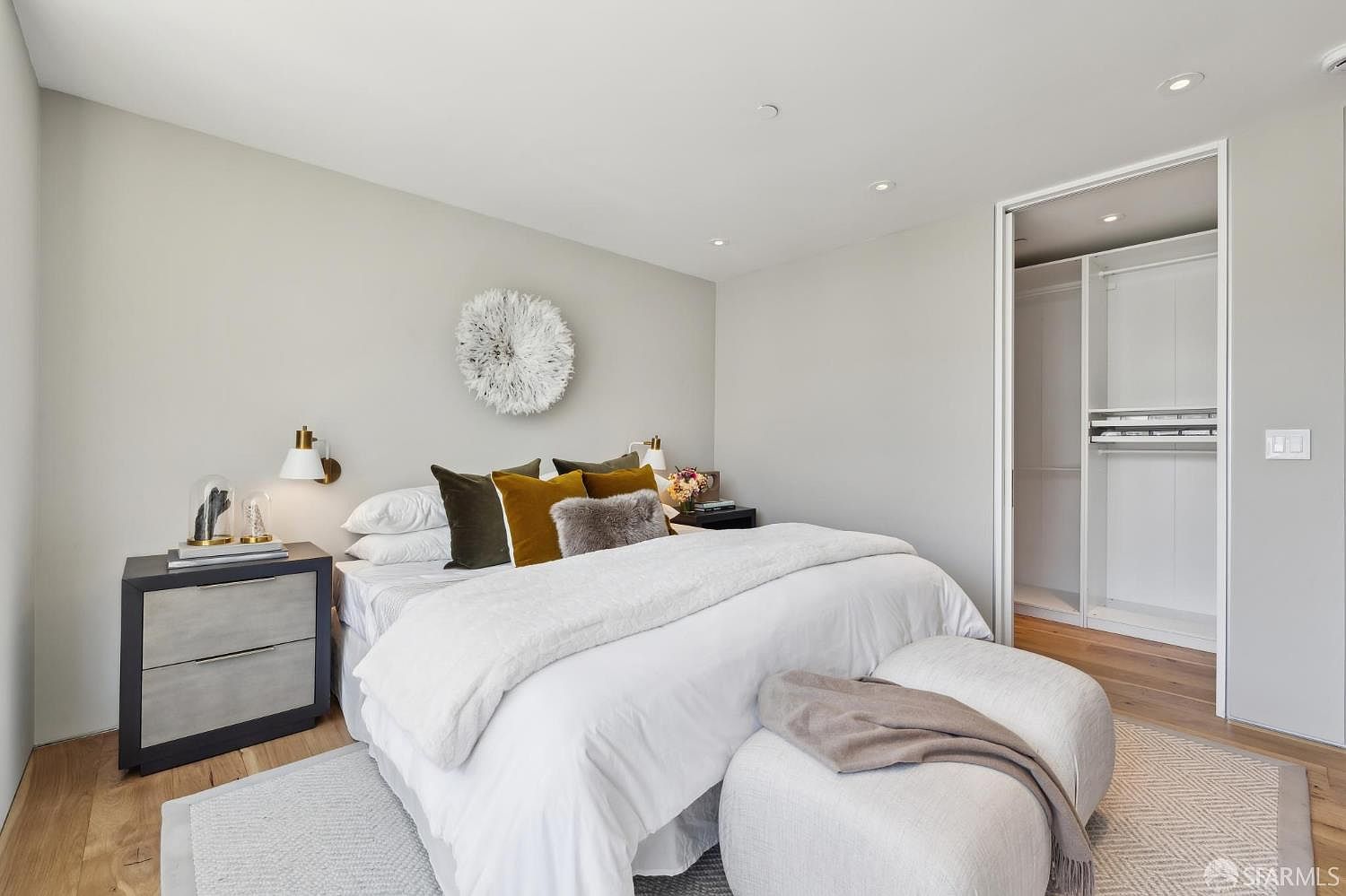 This bright and serene primary bedroom features a plush bed dressed in white linens, accented by a collection of olive and mustard-toned throw pillows and a textured decorative wall hanging. A modern nightstand with a sleek lamp sits to the left, while a large, open-concept walk-in closet with built-in shelving is visible through a doorway on the right. The room is finished with light wood flooring and a soft, neutral color palette, creating a calm and inviting atmosphere.