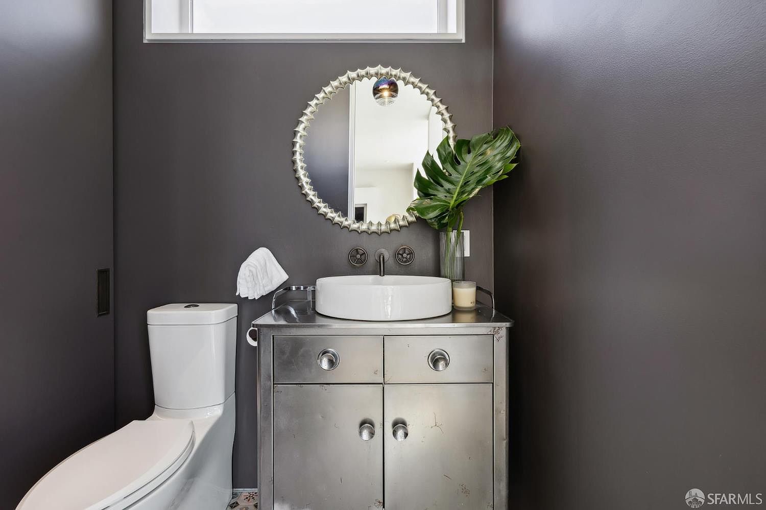 This modern powder room features a striking industrial aesthetic with dark charcoal walls and a metallic, stainless steel vanity cabinet. A circular, textured silver mirror hangs above a white vessel sink, complemented by a decorative vase with a large green leaf. The space is finished with a sleek white toilet and a transom window above, creating a bold and contemporary atmosphere.