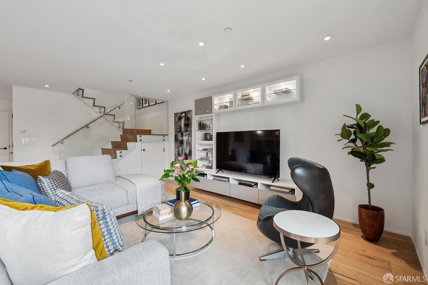 This bright and contemporary living room features a light-colored sectional sofa accented with blue and yellow pillows, paired with a modern dark-toned lounge chair. A sleek, white media console with integrated shelving and glass-front cabinets provides ample storage beneath a wall-mounted television. To the left, a staircase with glass railings and light wood treads adds an architectural element, while a potted plant and a glass-topped coffee table complete the inviting, open-concept space.