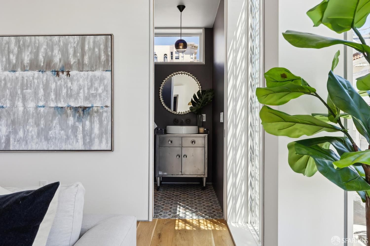 This view captures a modern powder room framed by a doorway, featuring a metallic vanity with a vessel sink and a circular decorative mirror. The space is accented by patterned floor tiles and a dark wall color, while a large abstract painting hangs on the adjacent white wall. To the right, a large plant sits near a window with a decorative screen, creating a bright and textured transition between the living area and the bathroom.