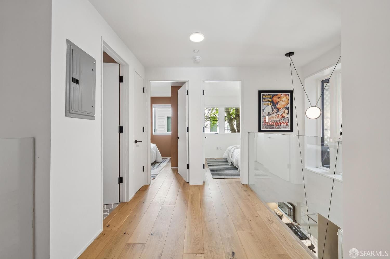 This bright, modern hallway features light wood flooring and crisp white walls, creating an airy and clean aesthetic. The space leads to two bedrooms and includes a glass-railed staircase overlooking the level below, accented by a contemporary pendant light and a framed movie poster. The perspective is centered, drawing the eye through the corridor toward the open doorways of the adjacent rooms.