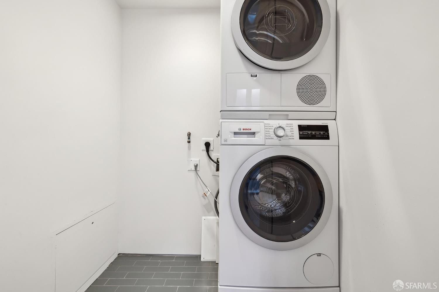 This compact laundry room features a stacked Bosch washer and dryer unit set against a clean, white-walled backdrop. The space is finished with grey rectangular floor tiles and includes visible utility connections on the adjacent wall. The minimalist design provides a functional and efficient laundry area.